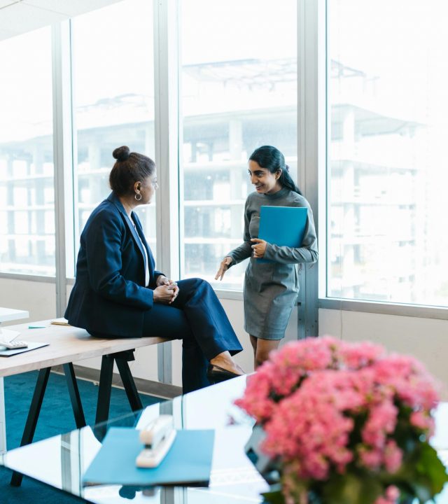 Two professional women in business attire having a conversation in a modern office setting.